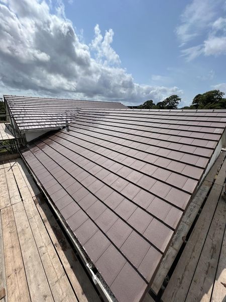 A newly installed brown tiled roof under a partly cloudy sky. The roof features uniform rectangular tiles, with wooden scaffolding surrounding the building's perimeter. Trees are visible in the distant landscape.