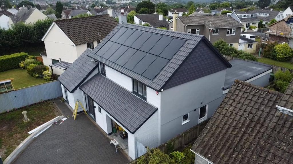 A modern, two-story house with a gray roof featuring solar panels. The exterior is primarily white with gray accents. The home is situated in a suburban neighborhood with other houses visible in the background.