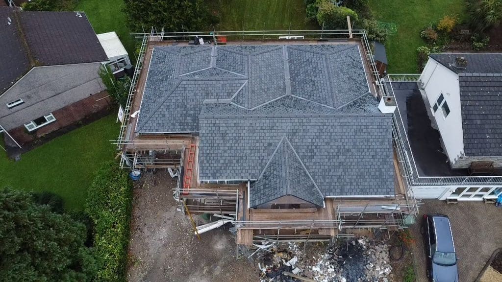 Aerial view of a house under construction with scaffoldings. The house has a complex roof design with multiple gables and gray shingles. There's a pile of construction debris in the front yard. The surrounding area is green with adjacent houses.