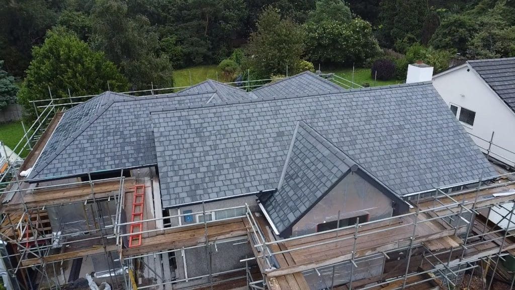 Aerial view of a house with a newly installed slate roof surrounded by scaffolding. The house is situated in a lush, green area with trees and grass in the background.