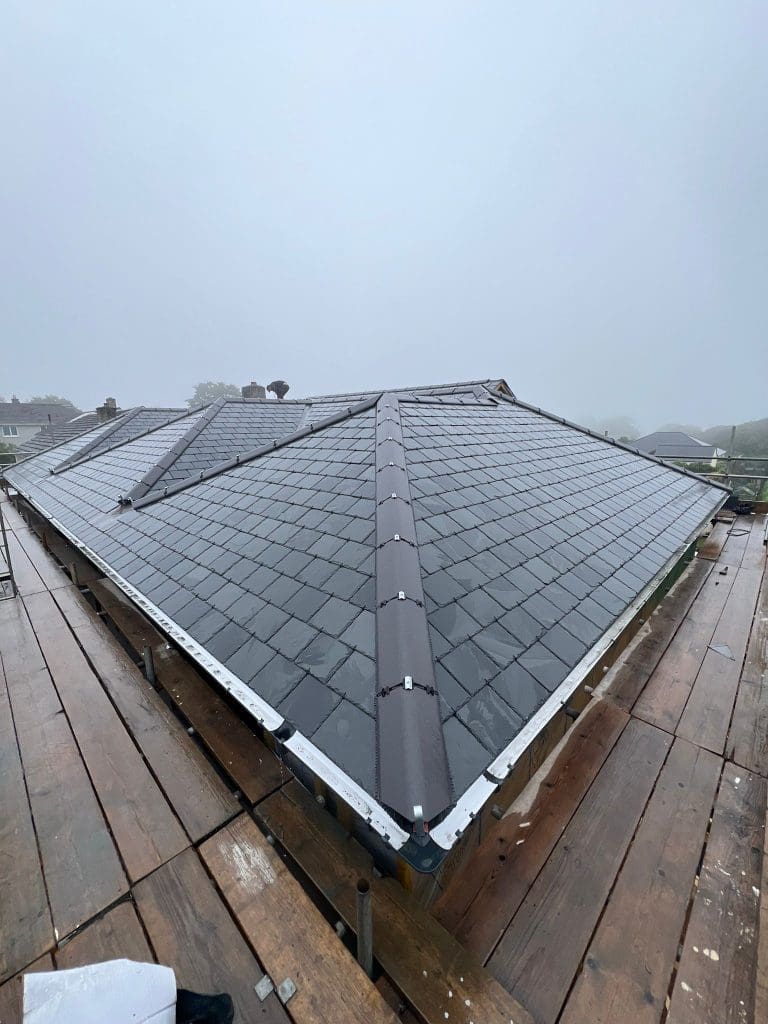 A new slate roof with a central ridge is shown on a misty day. The roof is surrounded by scaffolding planks, indicating ongoing construction work. Trees and houses are faintly visible in the foggy background.