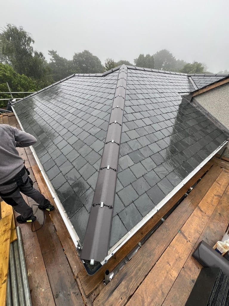 A person stands on a wooden platform next to a newly installed slate roof. The roof is wet and shiny, with dark gray tiles and black ridge caps. Trees and fog are visible in the background, indicating a misty day.