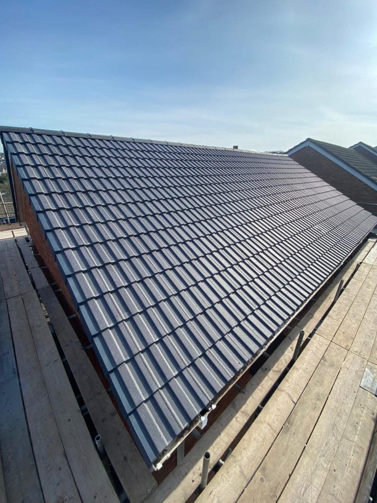 A new roof with dark gray tiles is shown under a clear blue sky. The structure is seen from a low angle, highlighting the symmetrical rows of tiles. Scaffolding surrounds the roof, indicating ongoing construction or renovation work.
