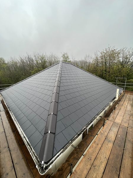 A triangular, newly installed roof with dark shingles and a central ridge viewed from a wooden platform. Surrounding trees hint at an overcast day.