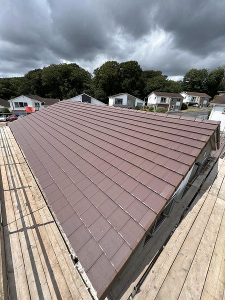 A roof with brown tiles on a residential building is shown. Wooden scaffolding surrounds the roof, and trees and cloudy skies are visible in the background. Several houses can be seen nearby.