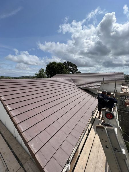 Two workers are standing on scaffolding while installing a roof with red tiles on a building under construction. The sky is partly cloudy, and trees are visible in the background.