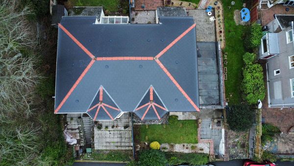 Aerial view of a large house with a dark gray roof and orange accents. The house is surrounded by a garden with greenery, pathways, and stone patios on both sides. A driveway with a red car is visible on the right.
