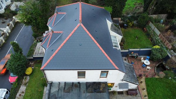 Aerial view of a house with a dark gray, pyramid-shaped roof featuring red accents along the edges. The property has a small front yard and a backyard with a brick patio and garden. A paved road runs alongside the house with passing cars.