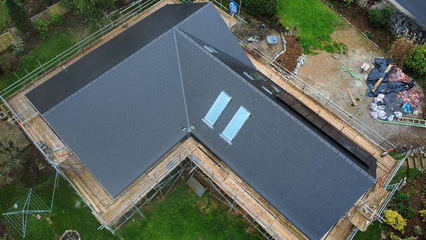 Aerial view of a house under construction with a newly installed dark gray roof. Two skylights are visible on one section. Scaffolding surrounds the house, and there are patches of dirt and grass in the surrounding yard.
