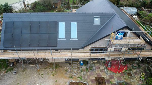 Aerial view of a house under construction with scaffolding. The roof has solar panels and skylights. A patio with various stones and a small garden are visible beside the house.