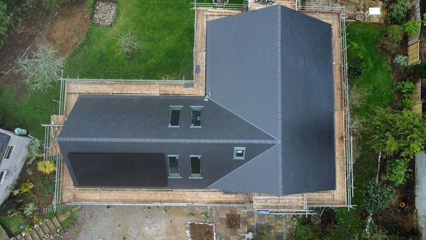 Aerial view of a house with a dark gray, polygonal roof surrounded by scaffolding. The house is set in a grassy area with some trees and bushes, and there is a paved area at the bottom.