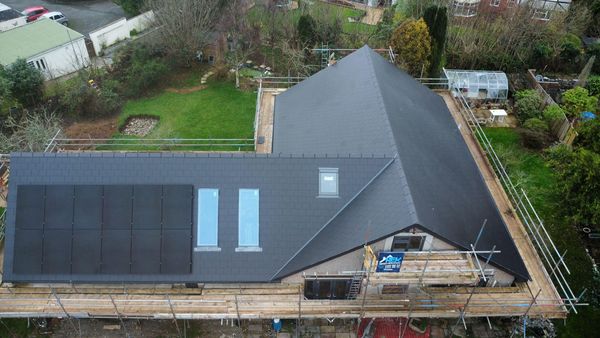 Aerial view of a house with a dark roof, solar panels, and skylights. Scaffolding surrounds the structure. The property is set in a green garden with trees, shrubs, and a greenhouse in the background.
