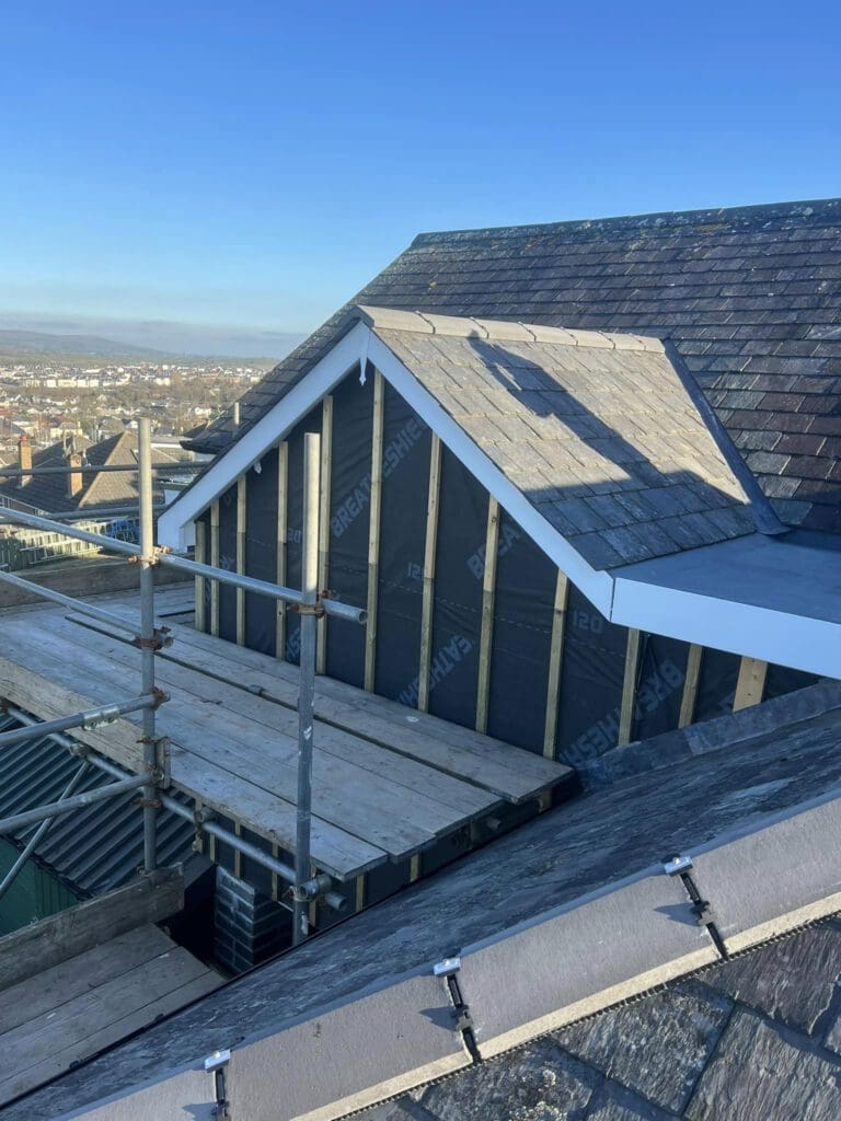 Rooftop under construction with wooden beams and partial slate tiling. Scaffolding is visible on the left side, with a clear blue sky and distant cityscape in the background.