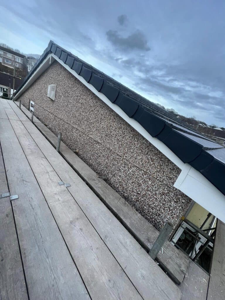 View of a roof on a cloudy day. The roof features dark shingles and a pebbledash exterior wall. Scaffolding is present alongside the building, indicating construction work. Some buildings are visible in the background.