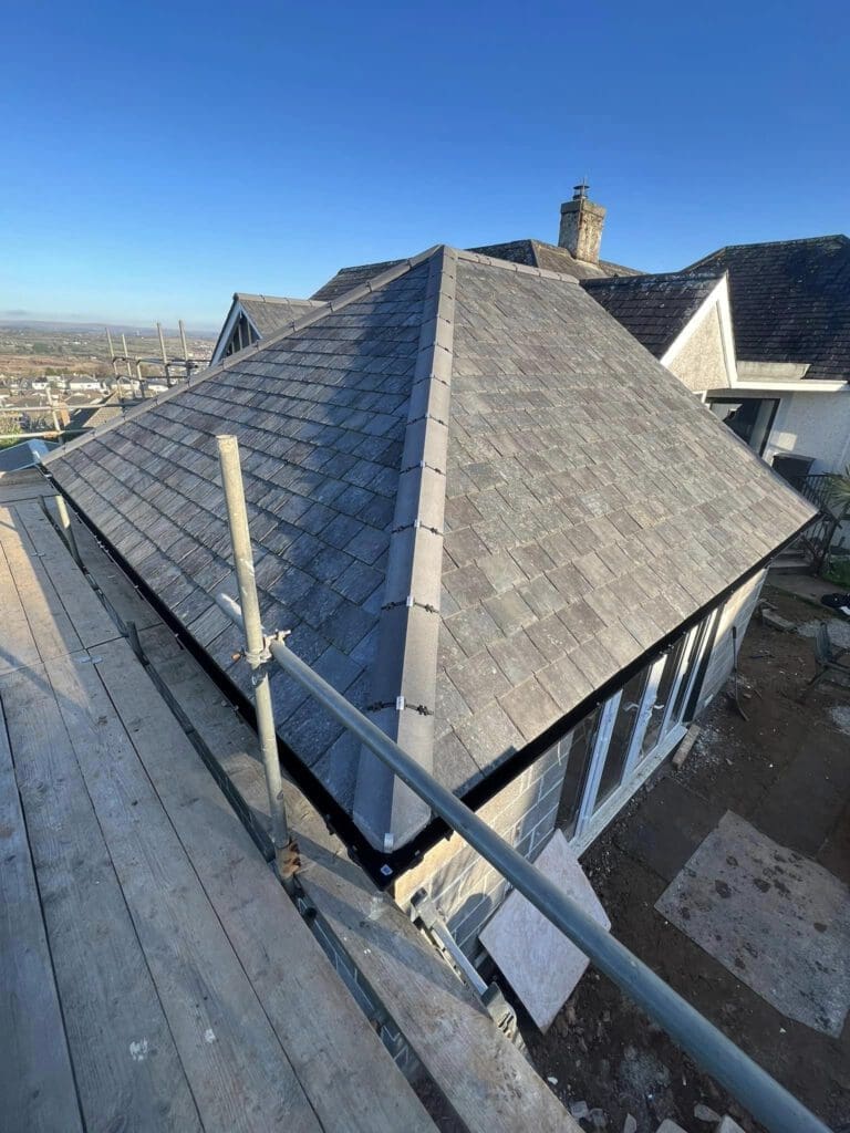 A newly constructed slate roof on a residential building with scaffolding around it. The sky is clear, and the landscape includes distant buildings and a chimney visible in the background.