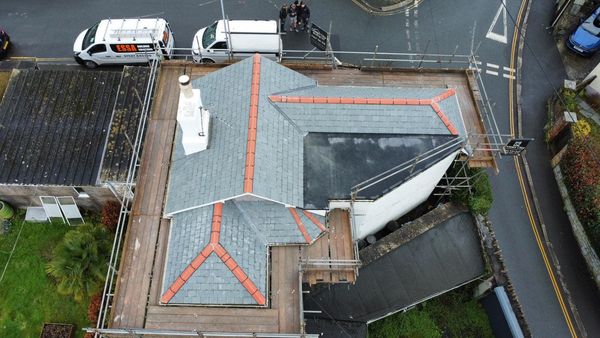 Aerial view of a building under construction with scaffolding around it. The structure has a complex, angular slate roof with red ridge tiles. Nearby are two vans parked on the street, and a group of people stands beside them, near an intersection.