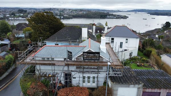 Aerial view of a house under construction, surrounded by scaffolding with a sign that reads "Ocean City." The house is in a residential area near a body of water, with other houses nearby and a scenic view of the waterfront and distant land.