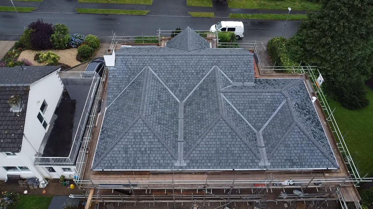 Aerial view of a house with a newly installed slate roof featuring multiple gables. The structure is surrounded by scaffolding, and construction materials are visible. A street and garden with greenery are in the background.